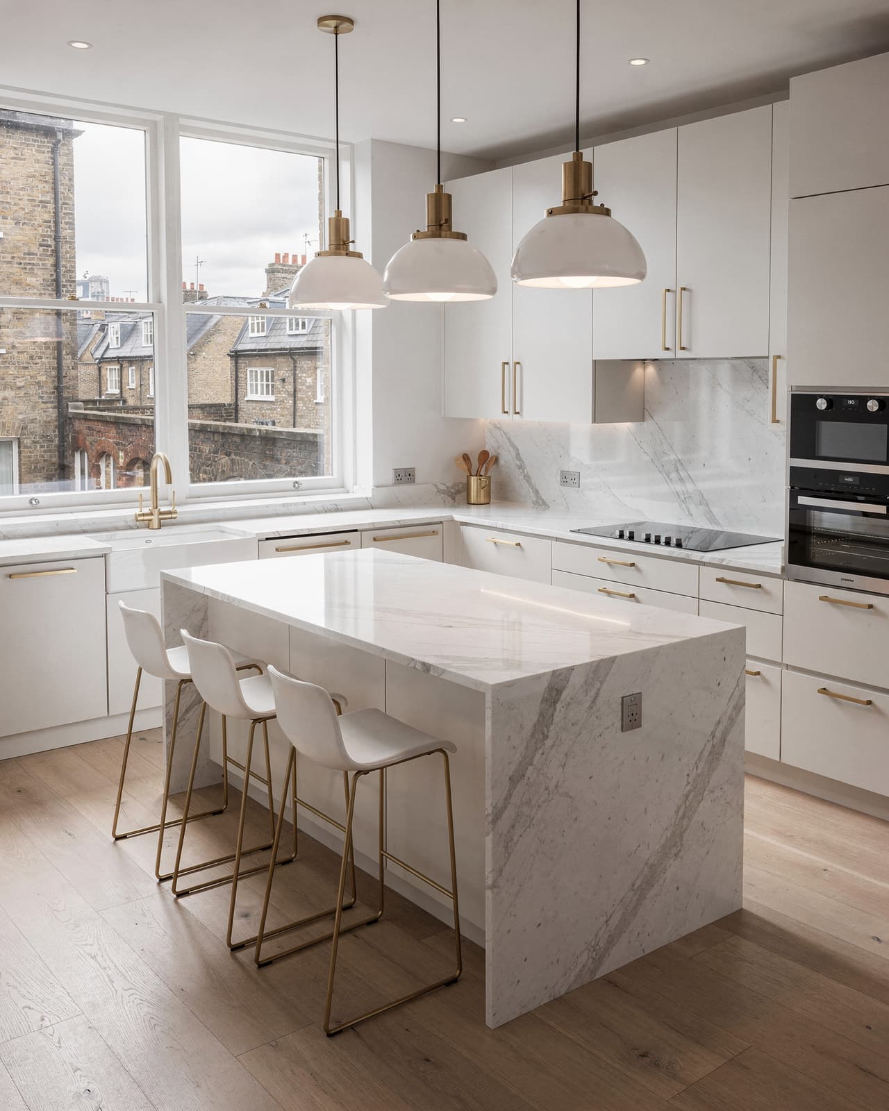 A bright, modern kitchen with white marble countertops, gold hardware, minimalist cabinetry, and large pendant lights over a central island